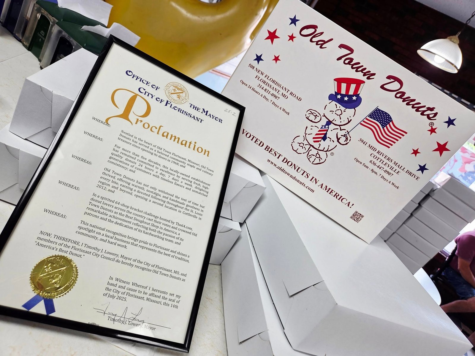 Framed City of Florissant proclamation declaring Old Town Donuts as "America's Best Donut," displayed next to a branded Old Town Donuts box with patriotic graphics and contact information.