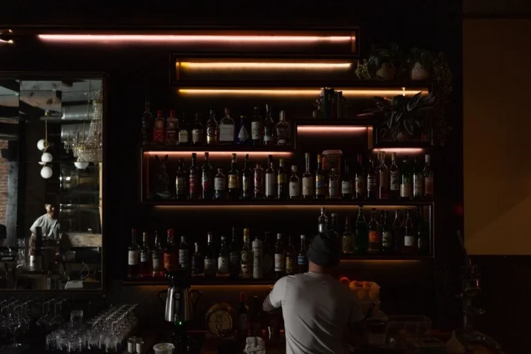 Bartender working in the moody, backlit bar at Savage & The Accomplice in St. Louis, with shelves of spirits and ambient lighting.