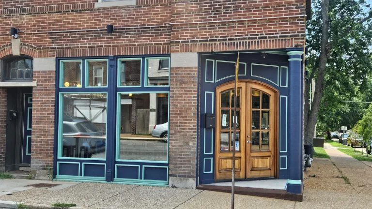 Charming exterior of a Tower Grove South restaurant featuring wooden double doors and teal-accented brickwork on a sunny corner