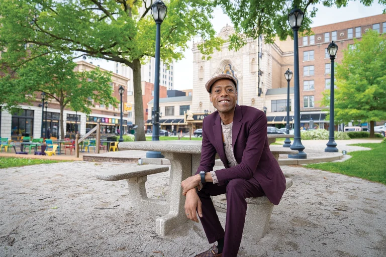 Tai Davis sitting at a stone picnic table in Strauss Park, with The Fabulous Fox Theatre and colorful Grand Center storefronts in the background, St. Louis, MO.